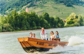 Gruppe von Menschen auf einem Holzboot auf der Donau mit grüner Landschaft im Hintergrund.