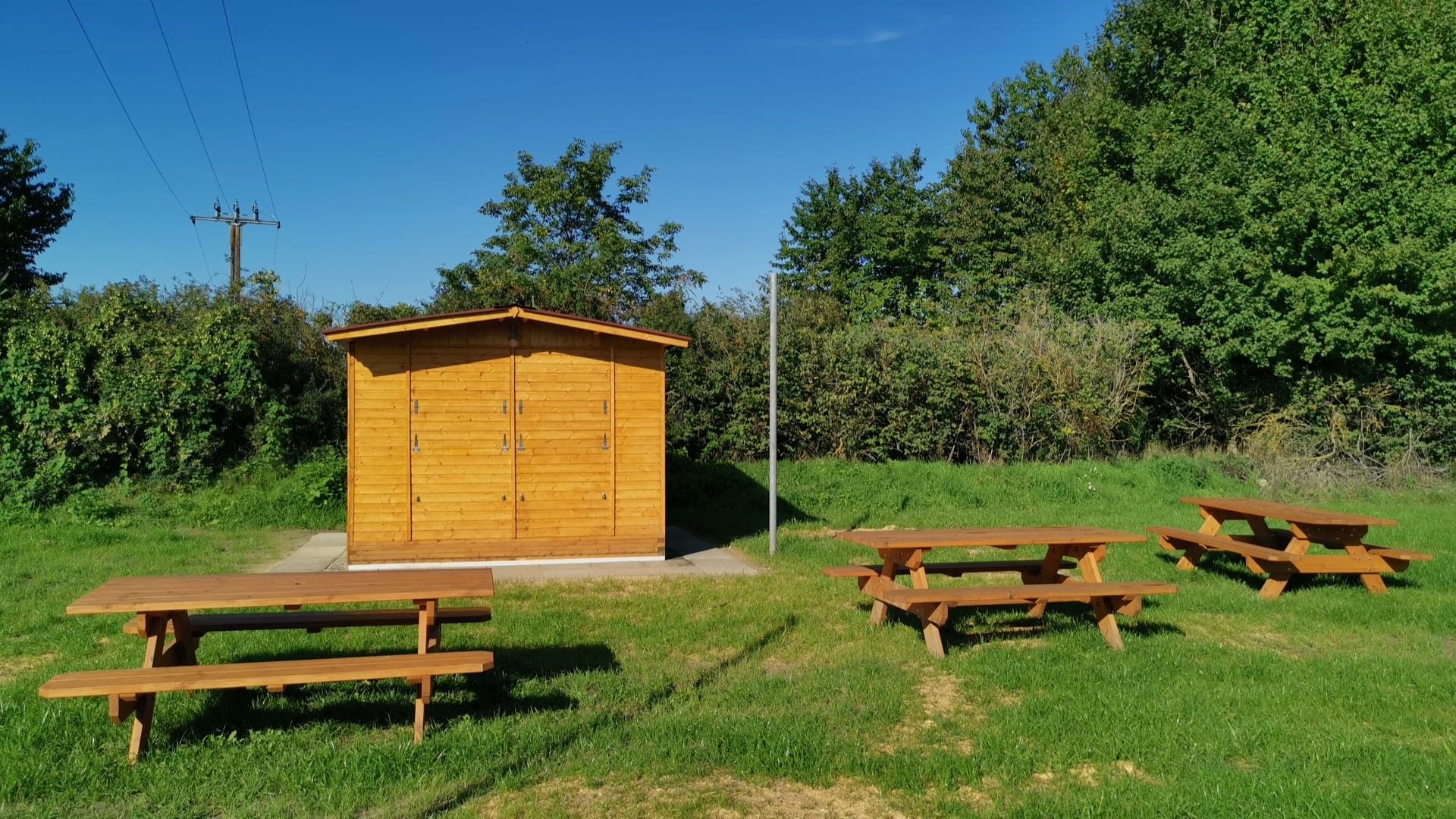 Holzhütte mit Picknicktischen auf einer Wiese, umgeben von Bäumen und Sträuchern unter blauem Himmel.