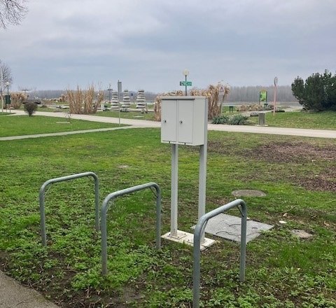 E-Bike Ladestation auf einer Wiese mit Fahrradst&auml;ndern und einem Weg im Hintergrund.