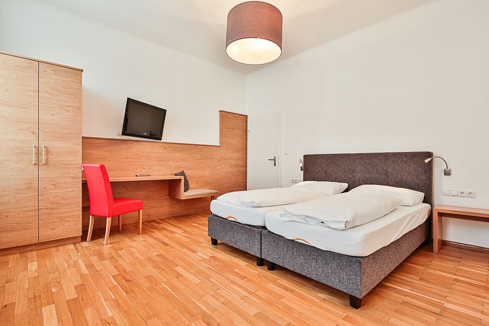 Hotelzimmer mit Doppelbett, rotem Stuhl, Schreibtisch und Fernseher.