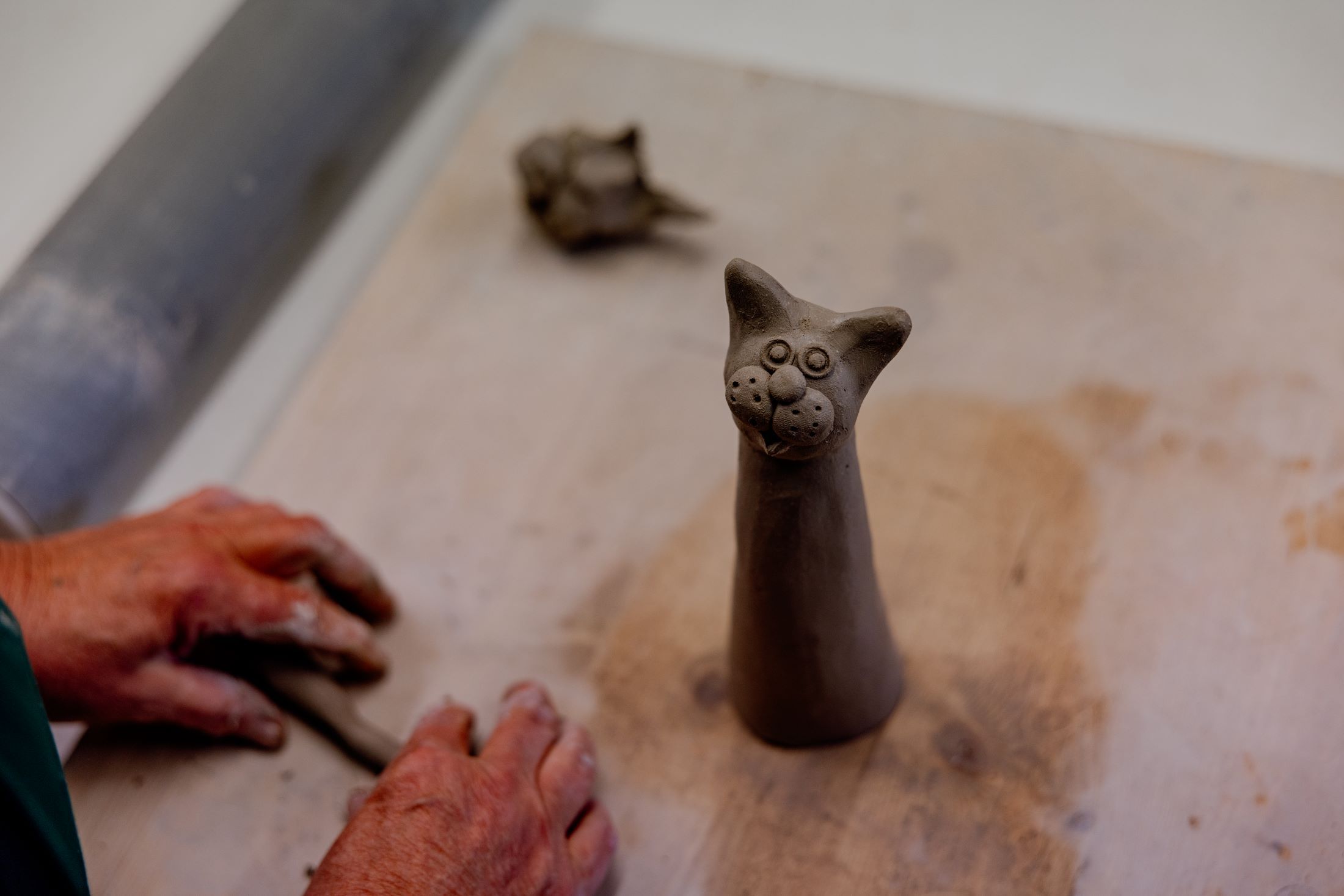 Eine handgefertigte Tonfigur einer Katze auf einem Tisch, daneben Hände, die Ton formen.