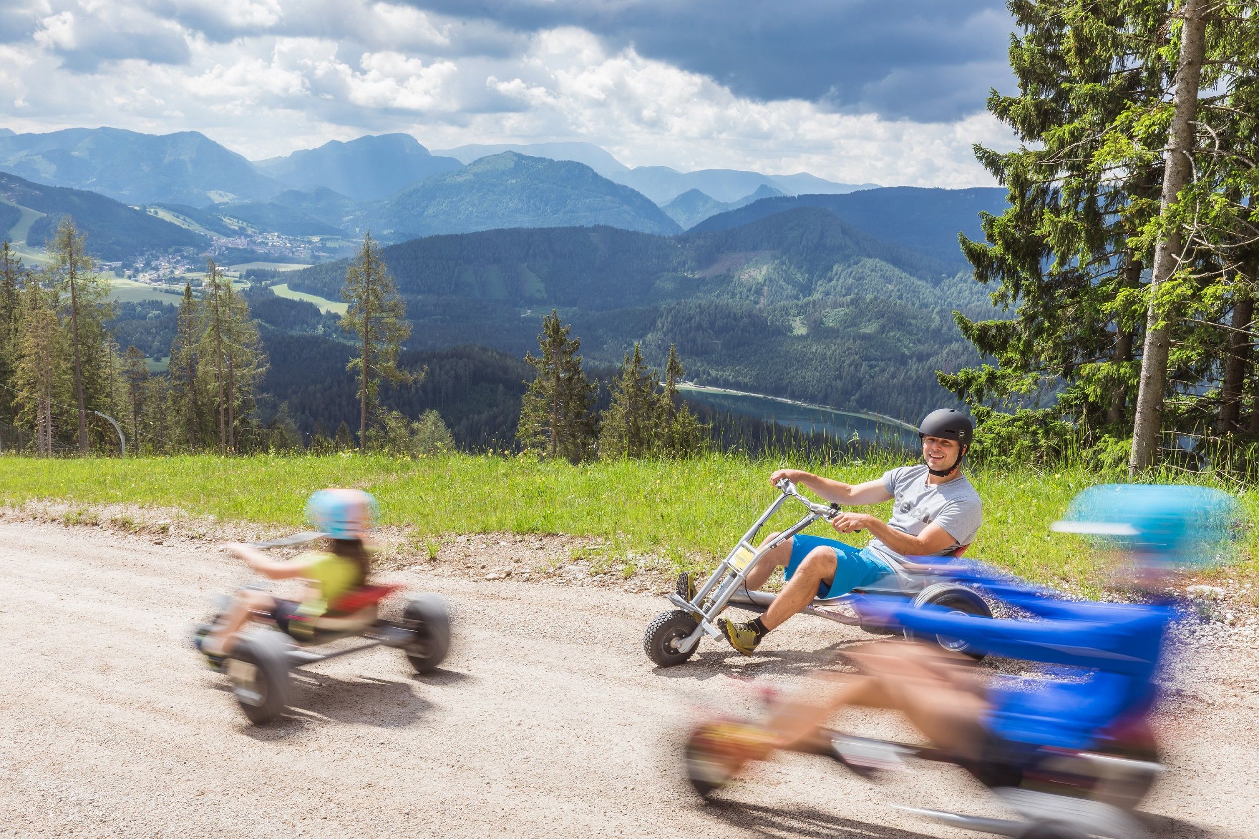 Personen fahren auf Dreirädern einen Hügel hinunter mit Berglandschaft im Hintergrund.