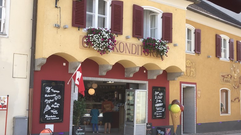 Au&szlig;enansicht eines Caf&eacute;s mit gelber Fassade, roten Fensterl&auml;den und Blumendekoration.