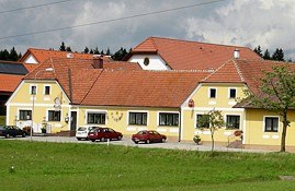 Café-Restaurant Schrammel**, © Café-Restaurant Schrammel** Gelbes Gebäude mit roten Dächern und mehreren Autos davor, umgeben von grüner Wiese und Bäumen.