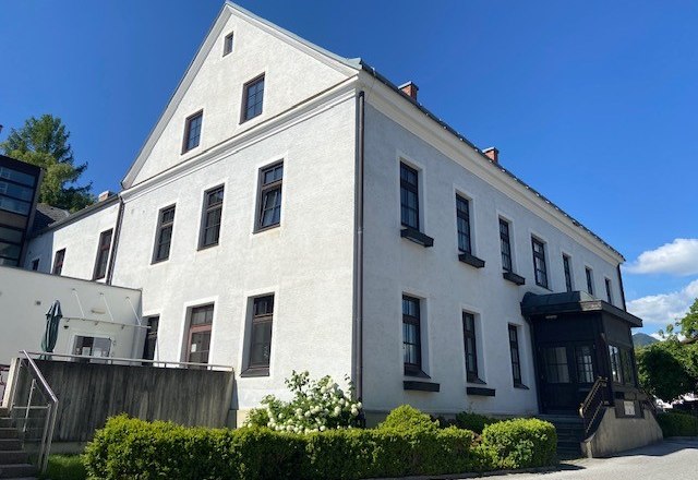 Ein gro&szlig;es, wei&szlig;es Geb&auml;ude mit mehreren Fenstern und einem Giebeldach, umgeben von gr&uuml;nen B&uuml;schen und einem blauen Himmel im Hintergrund.