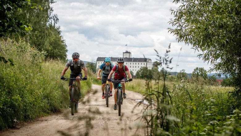 Drei Mountainbiker fahren auf einem Feldweg, im Hintergrund ein Schloss.