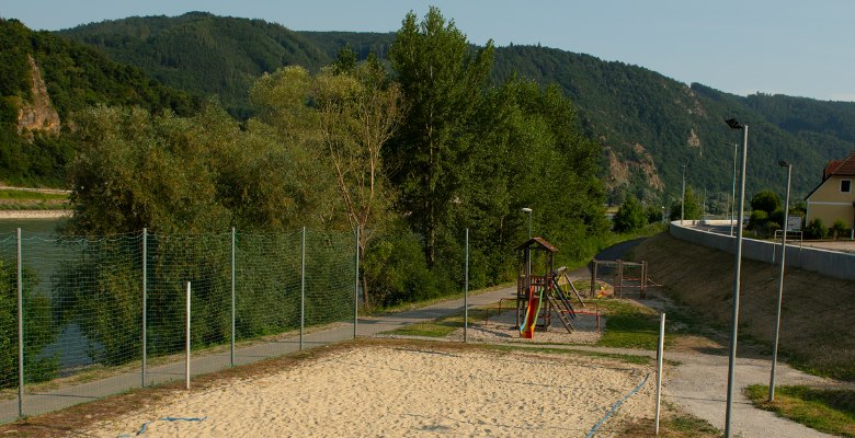 Ein Beachvolleyballplatz neben der Donau mit umliegenden Bäumen und einem Spielplatz im Hintergrund.