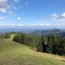 Blick auf eine grüne Berglandschaft mit Wäldern und blauem Himmel.