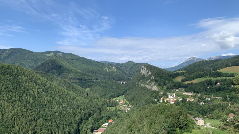 Panoramablick auf eine gr&uuml;ne Berglandschaft mit einem kleinen Dorf und einem Viadukt.