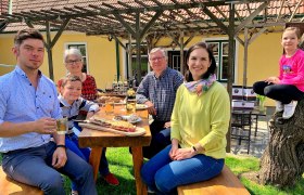 Eine Familie sitzt an einem Holztisch im Freien und genießt eine Mahlzeit. Ein Kind sitzt auf einem Baumstamm. Im Hintergrund ein gelbes Haus mit Pergola.