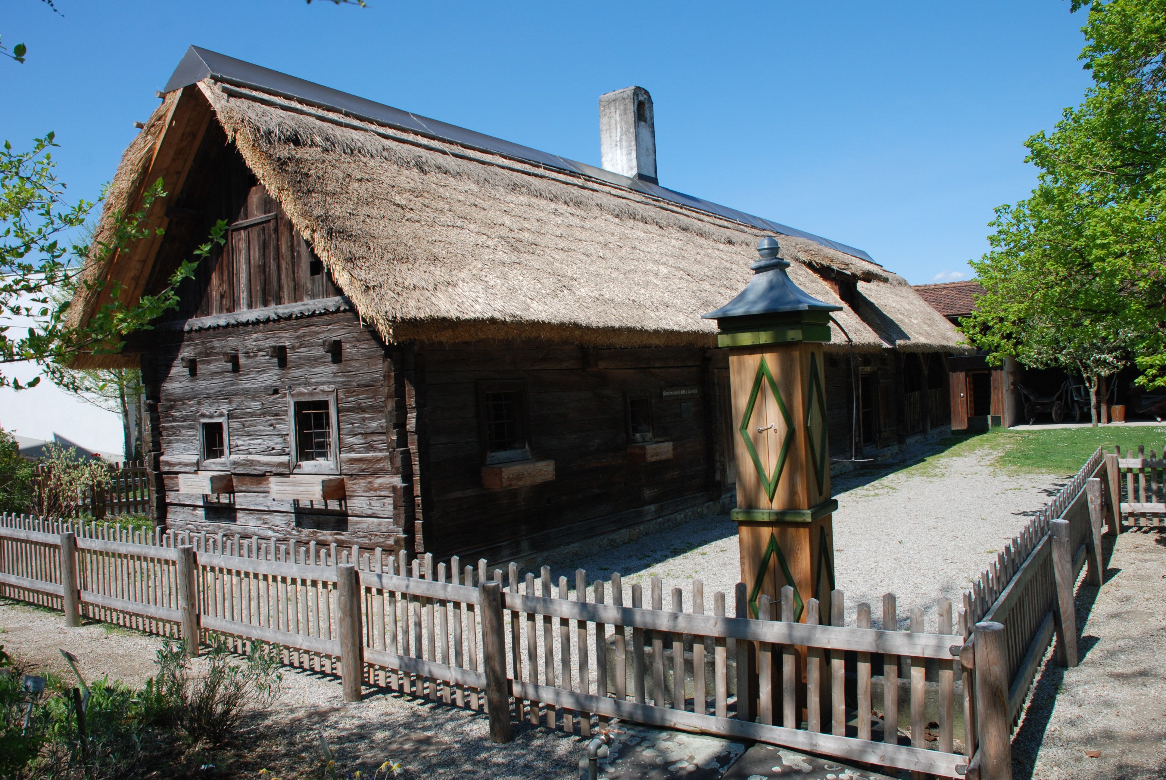 Traditionelles Holzhaus mit Strohdach und Holzzaun in ländlicher Umgebung.