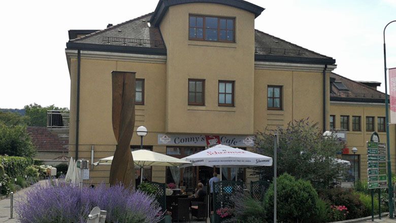 Außenansicht von Conny's Café in Gars mit Sonnenschirmen und Pflanzen.