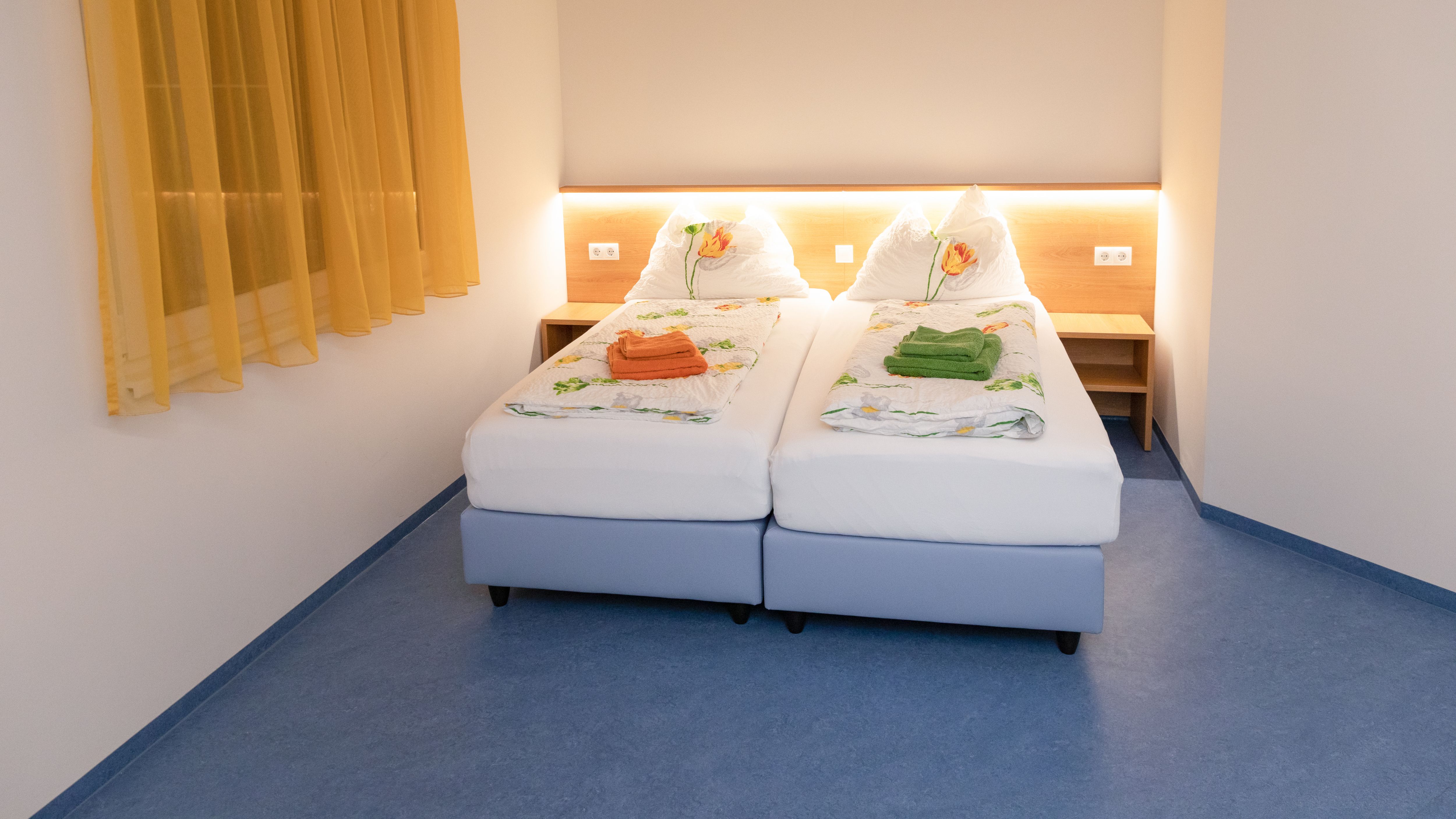 Ein Doppelzimmer mit zwei Einzelbetten, gelben Vorhängen und blauer Boden. Die Betten sind mit Blumenbettwäsche und farbigen Handtüchern dekoriert.