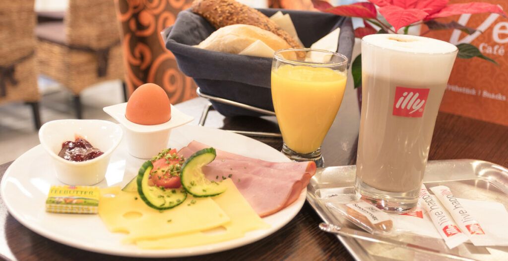 Ein Frühstücksteller mit Käse, Schinken, Ei, Marmelade, Butter, Brötchen, Orangensaft und einem Latte Macchiato.