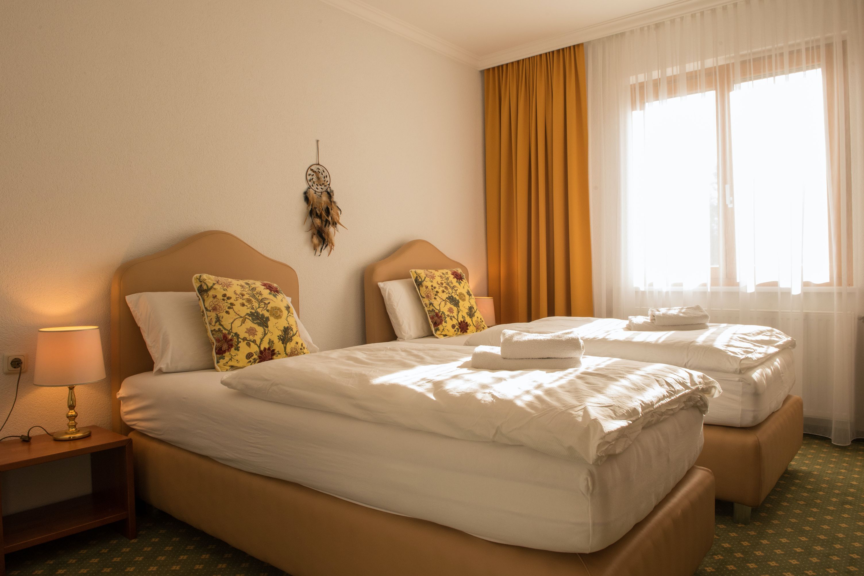 Ein gemütliches Doppelzimmer mit zwei Einzelbetten, gelben Vorhängen und einem Traumfänger an der Wand.