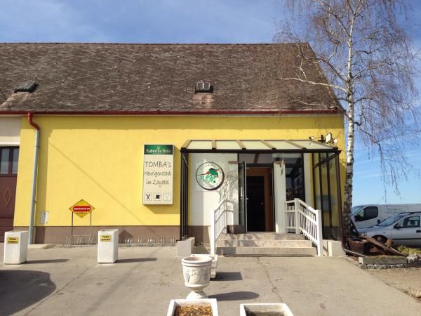 Gelbes Gebäude mit Schild 'Tomba's Heurigenschenke'. Eingang mit Treppe und Glasüberdachung.