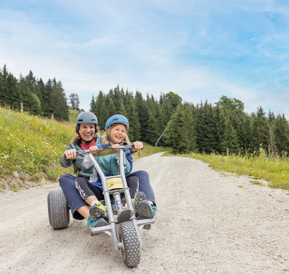 Zwei Personen fahren auf einem Mountaincart auf einem Schotterweg, umgeben von Wiesen und Wald.
