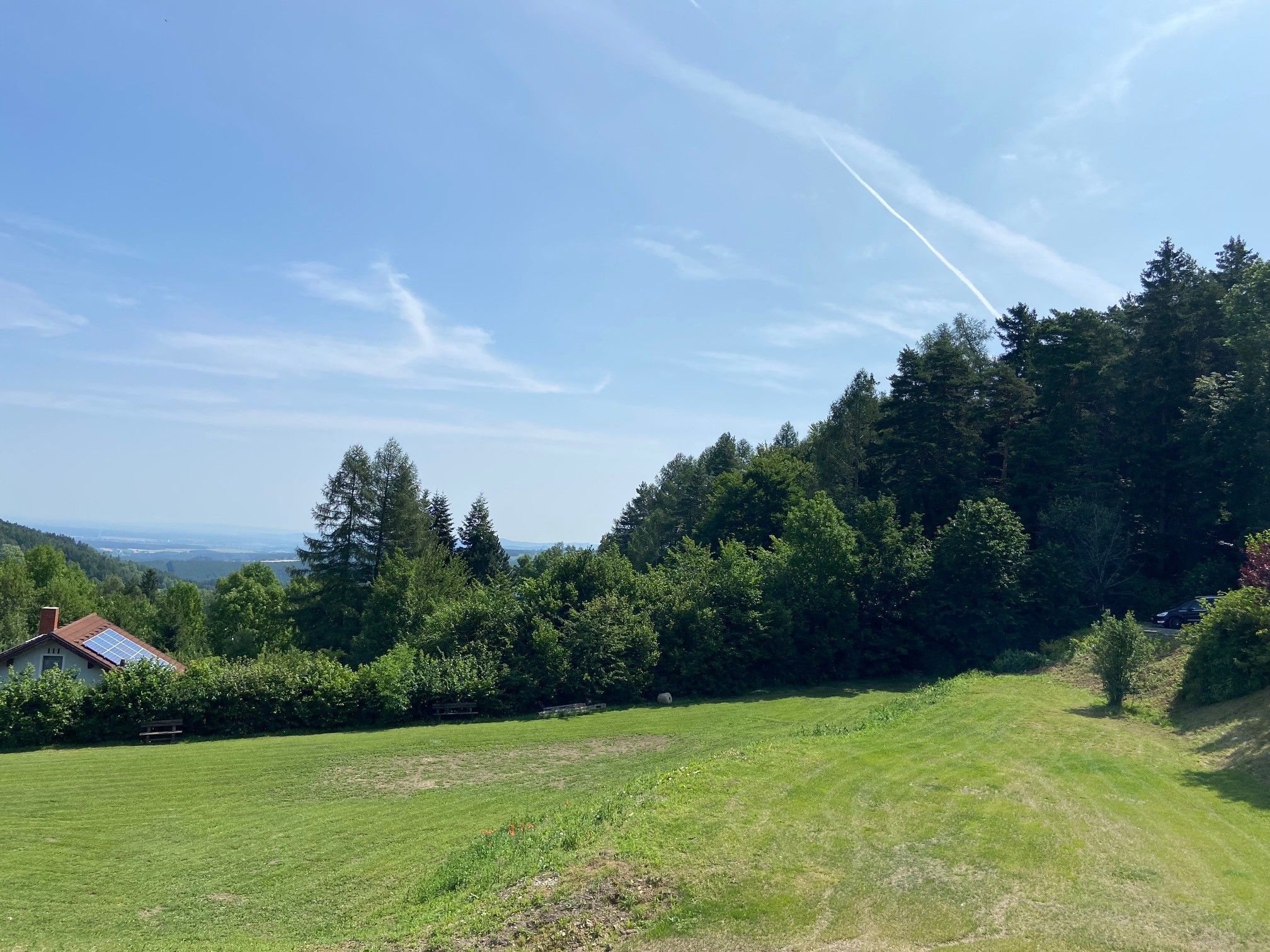 Grüne Wiese mit Bäumen und einem Haus mit Solarpanelen, blauer Himmel im Hintergrund.