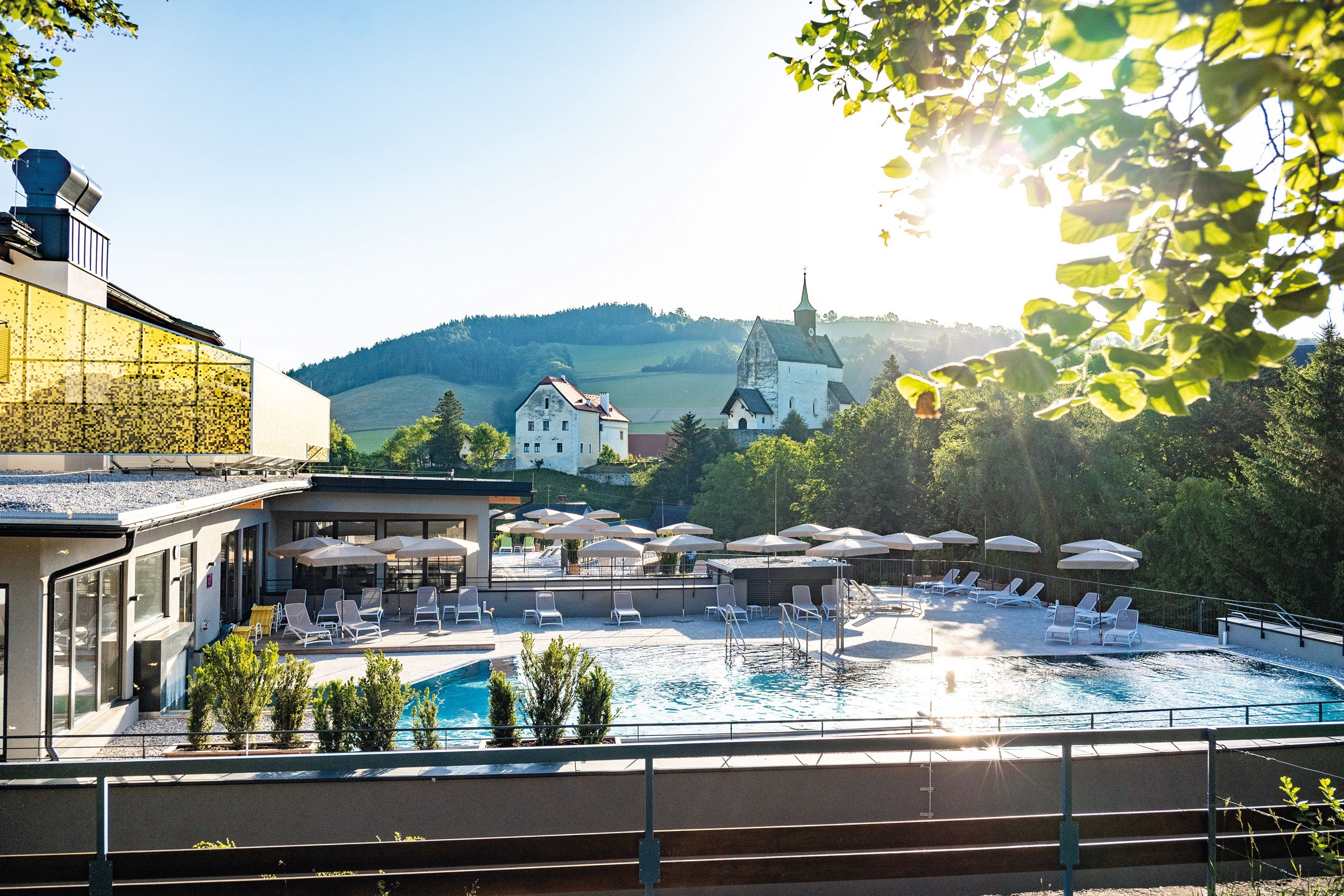 Außengelände eines Hotels mit Pool, Sonnenschirmen und Liegen, umgeben von Natur und Kirche im Hintergrund.