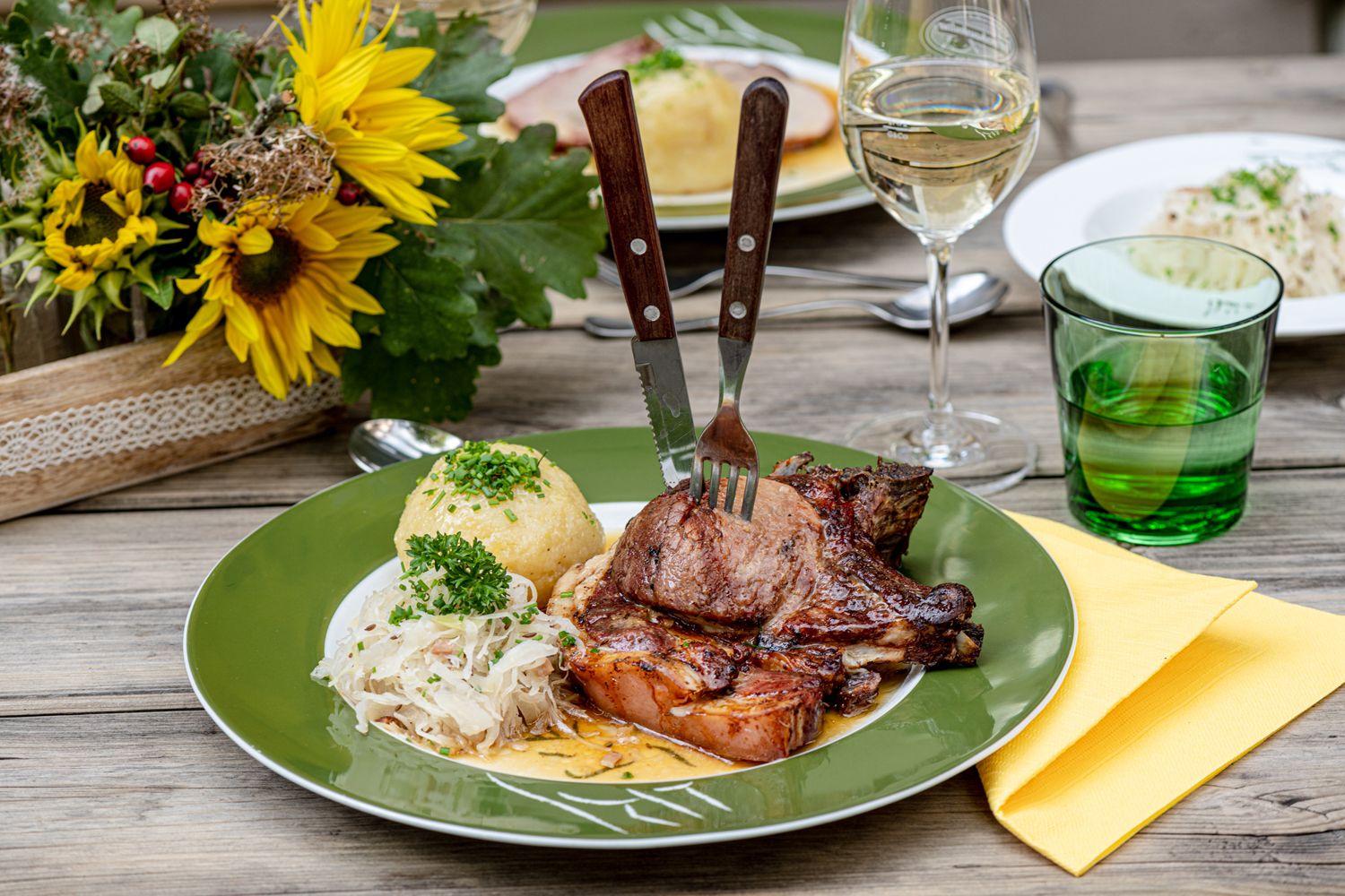 Ein Teller mit Schweinsbraten, Knödel und Sauerkraut auf einem Holztisch, daneben ein Glas Weißwein und ein Blumenarrangement.