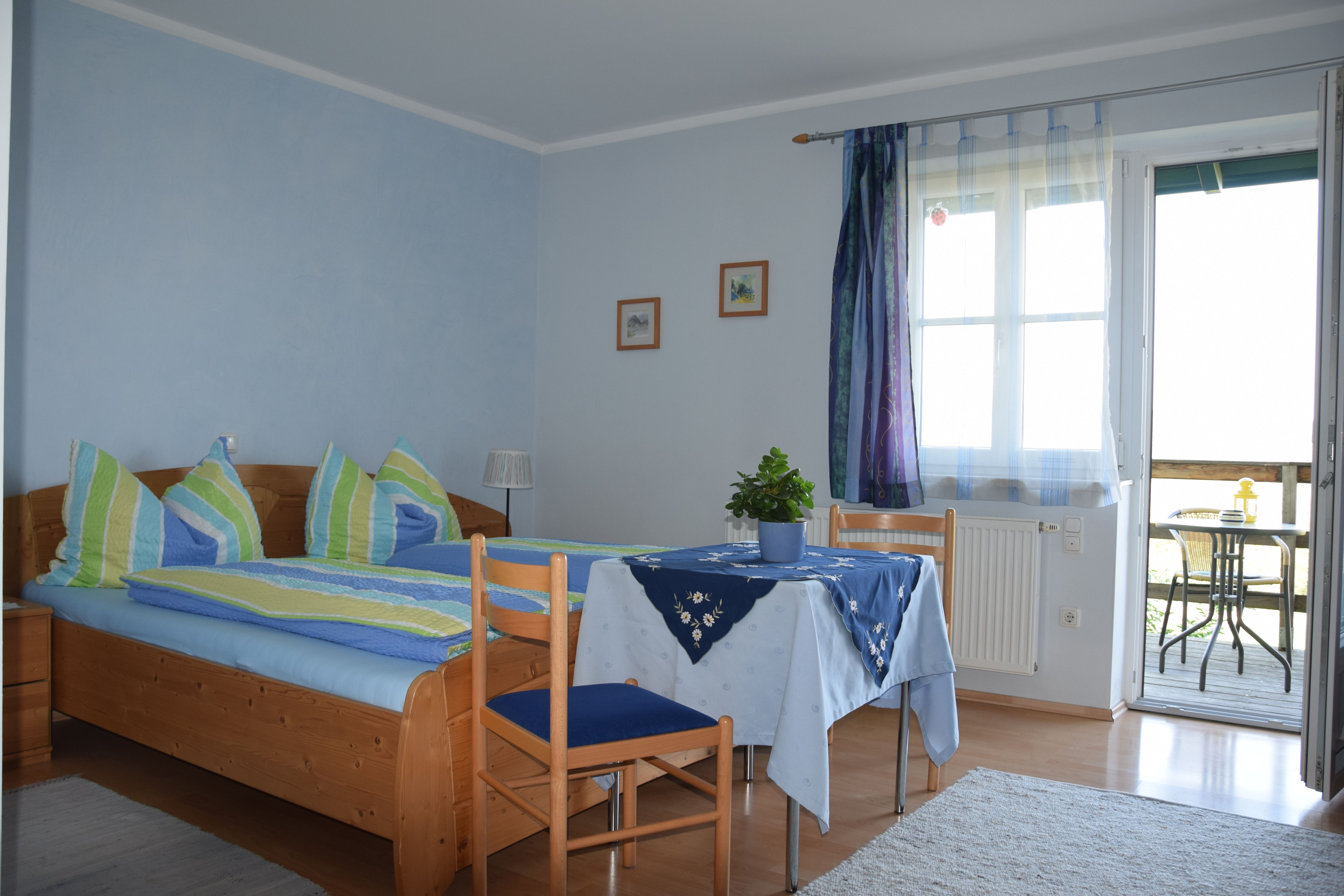 Gemütliches Gästezimmer mit Doppelbett, Tisch und Stühlen, blauen Vorhängen und Zugang zu einem Balkon mit Sitzgelegenheit.