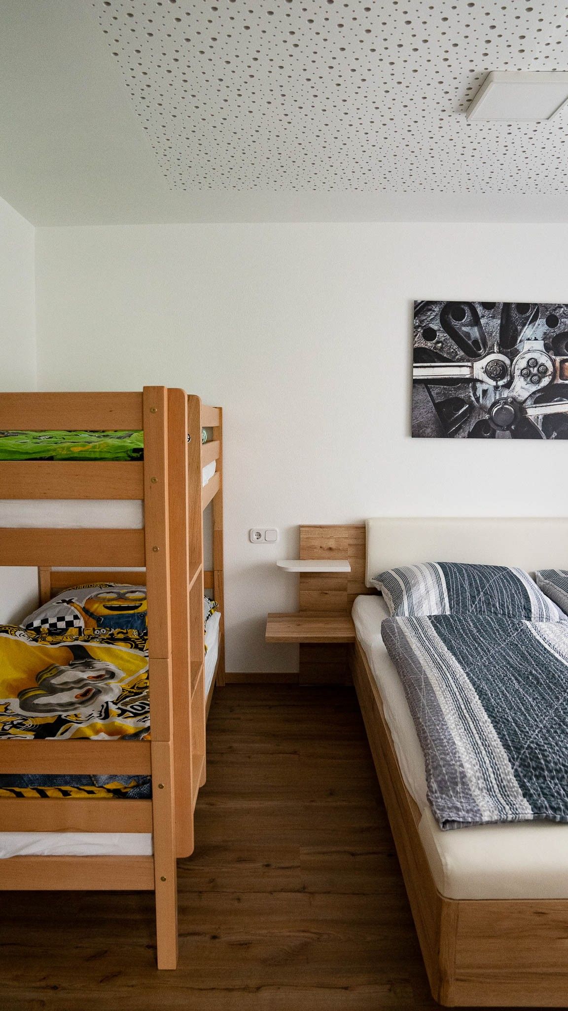 Ein Schlafzimmer mit einem Etagenbett und einem Doppelbett aus Holz. An der Wand hängt ein Bild mit einem technischen Motiv.