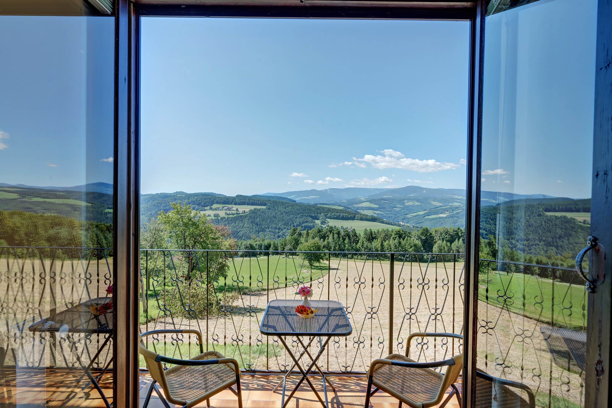 Blick von einem Balkon auf eine hügelige Landschaft mit Wäldern und Feldern, zwei Stühle und ein Tisch mit Blumen im Vordergrund.