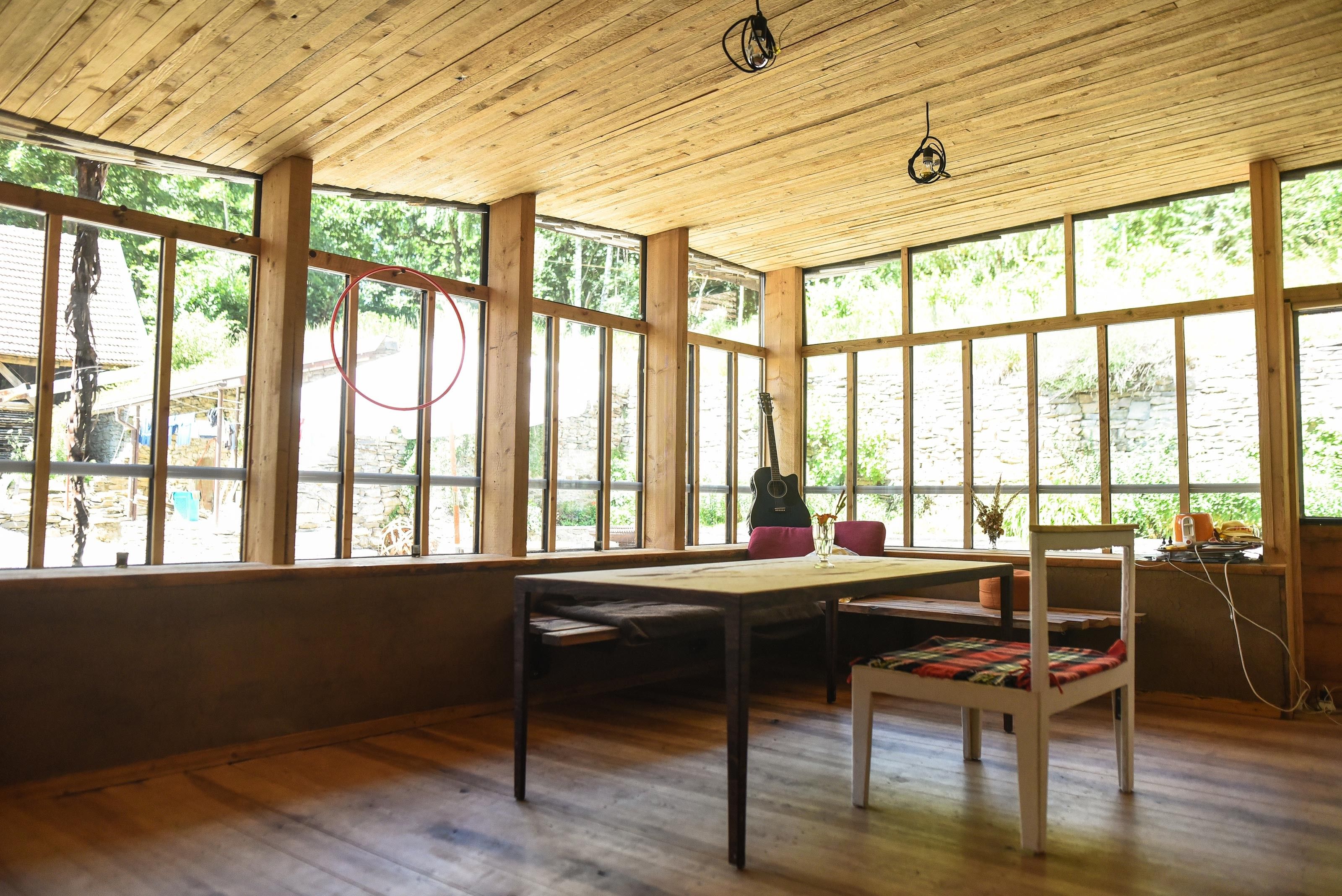 Ein gemütlicher Raum mit Holzboden und -decke, großen Fenstern, einem Tisch, Stühlen und einer Gitarre in der Ecke.