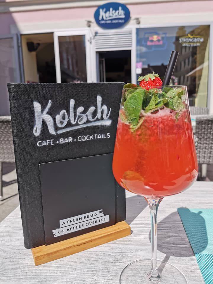 Ein roter Cocktail mit Minze und Erdbeere vor einem Café mit Schild 'Kölsch Café Bar Cocktails'.