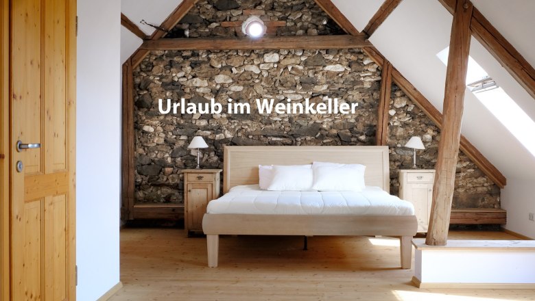 Schlafzimmer mit Holzbalken und Steinwand, Bett in der Mitte, Text 'Urlaub im Weinkeller'.