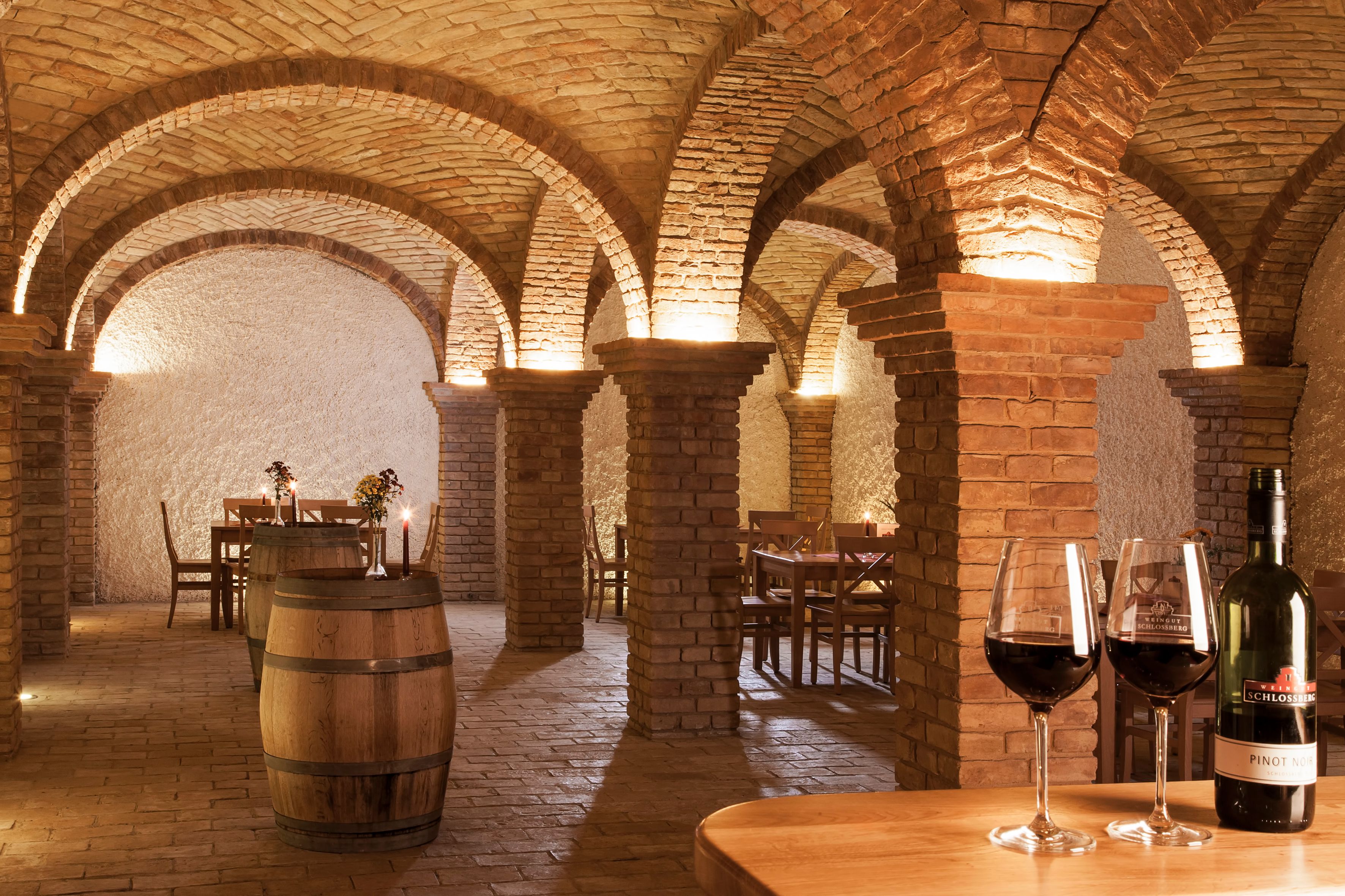 Weinkeller mit Backsteinbögen, Tischen und Weinfässern, beleuchtet durch Kerzen und Lampen.