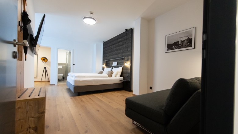 Modern eingerichtetes Hotelzimmer mit Doppelbett, Sofa und Flachbildfernseher. Helle Holzb&ouml;den und ein Badezimmer im Hintergrund.