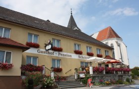 Ein traditioneller Gasthof mit Blumen geschmückter Fassade und Terrasse, daneben eine Kirche mit spitzem Turm.