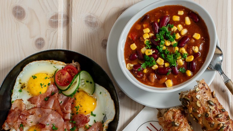 Verschiedene warme Snacks auf einem Tisch, darunter Sandwiches, Spiegelei mit Speck, Suppe und Br&ouml;tchen.