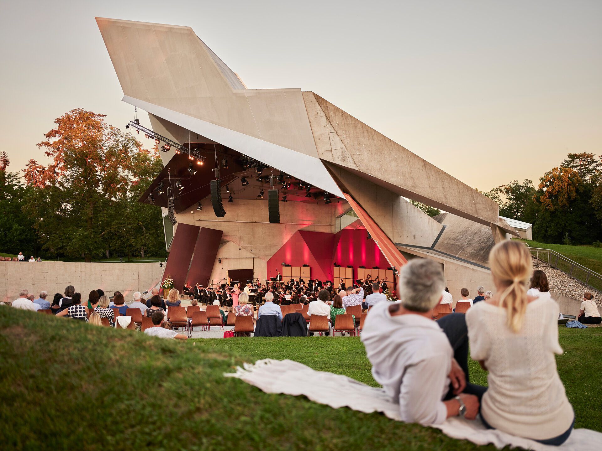 Ein lauer Sommerabend lädt dazu ein, die Klänge eines Konzerts unter freiem Himmel zu genießen. Die beeindruckende Architektur der Bühne harmoniert perfekt mit der umgebenden Natur, während Besucher auf Decken sitzen und die Atmosphäre in vollen Zügen auskosten.