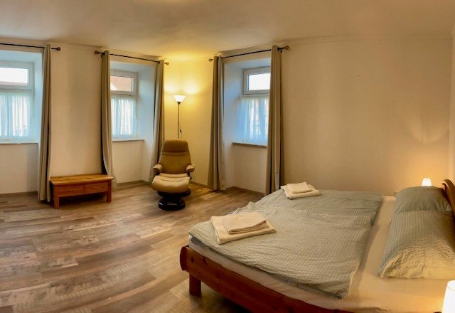 Ein schlichtes Schlafzimmer mit Doppelbett, Sessel, Nachttisch und drei Fenstern mit Vorh&auml;ngen.