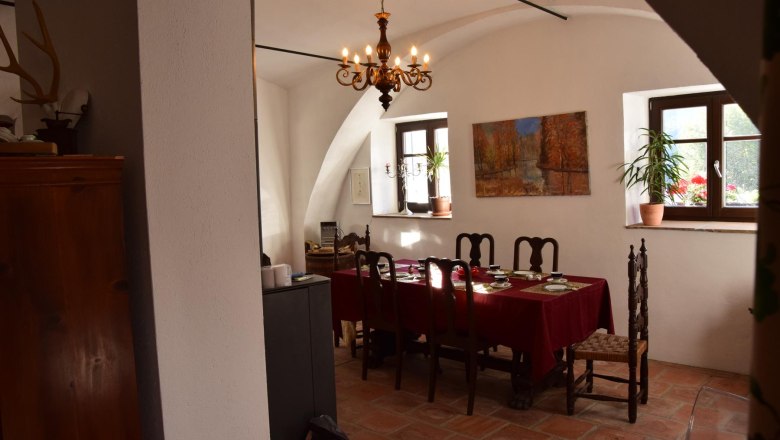 Ein Esszimmer mit einem Tisch, der mit einer roten Tischdecke gedeckt ist. An der Wand hängt ein Gemälde, und ein Kronleuchter hängt von der Decke.