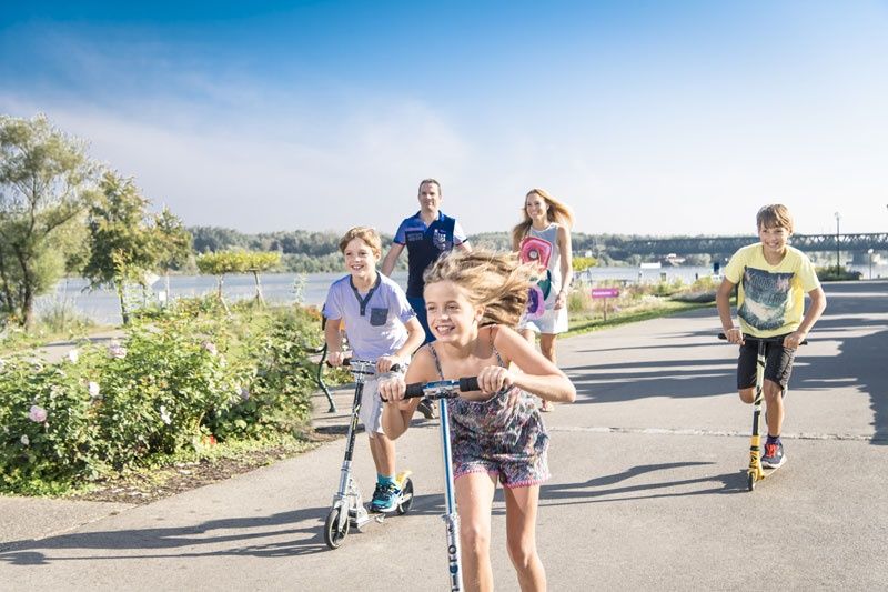 Kinder fahren mit Rollern auf einem Weg entlang eines Flusses, begleitet von zwei Erwachsenen im Hintergrund.