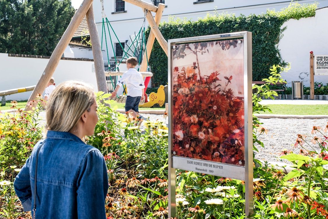 Eine Frau betrachtet ein Kunstwerk im Freien vor einem Museum, umgeben von Blumen und einem Spielplatz.