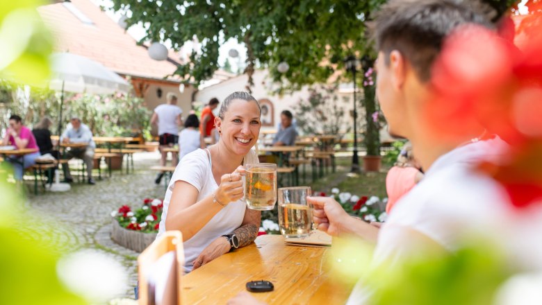 Personen sto&szlig;en mit Getr&auml;nken im Freien an, umgeben von B&auml;umen und Blumen.