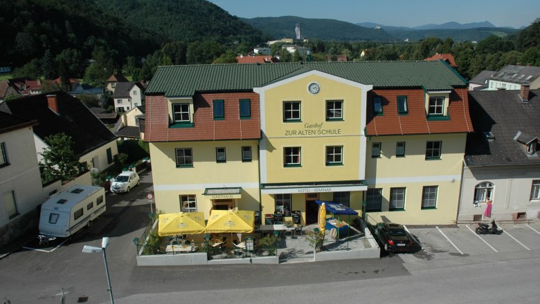Hotel Zur Alten Schule, © Viregro Capital Gmbh Gelbes Gebäude mit der Aufschrift 'Gasthof Zur Alten Schule', umgeben von Hügeln und Bäumen.