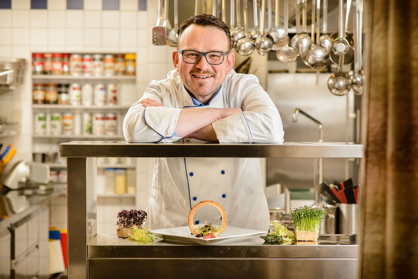 Ein Koch in weißer Uniform lehnt sich auf eine Edelstahltheke in einer Küche. Vor ihm steht ein Teller mit dekorativem Essen.