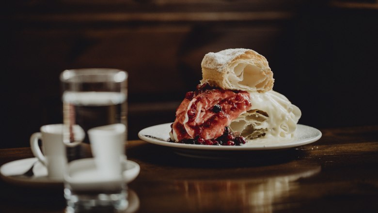 Ein Teller mit einer Klostercremeschnitte, garniert mit roten Beeren, steht auf einem Holztisch. Im Hintergrund sind ein Glas Wasser und zwei Espressotassen zu sehen.