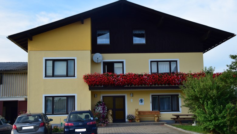 Au&szlig;enansicht eines gelben Hauses mit Balkon voller roter Blumen und geparkten Autos davor.