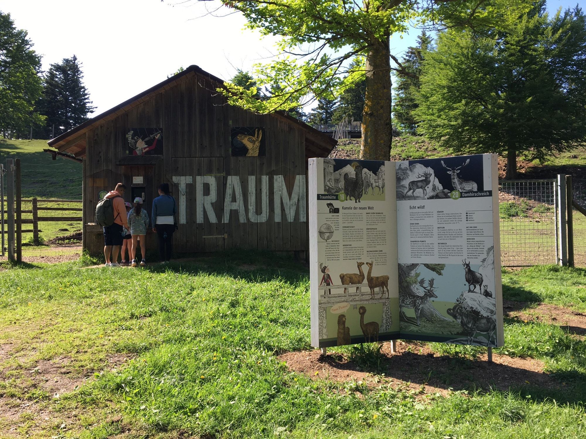 Eine Familie steht vor einer Holzhütte mit der Aufschrift 'TRAUM'. Daneben eine Infotafel über Tiere in einem grünen Park.