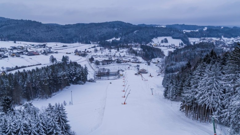 Verschneite Skipiste in Kirchbach, umgeben von W&auml;ldern und H&uuml;geln.