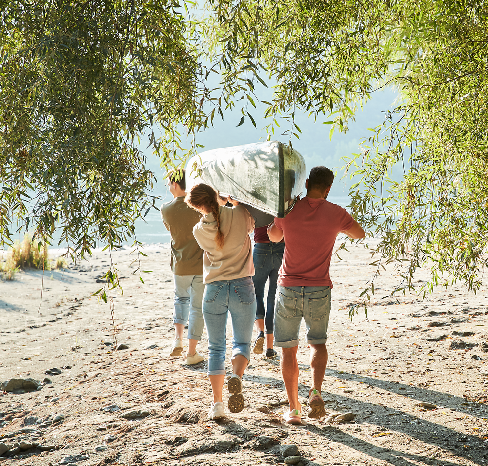 Eine Gruppe von Freunden genießt einen sonnigen Tag am Ufer, während sie ein Kanu tragen. Die sanften Wellen des Wassers und das Spiel von Licht und Schatten unter den Bäumen schaffen eine einladende Atmosphäre für Abenteuer und Entspannung. Hier wird der Sommer in vollen Zügen ausgelebt.