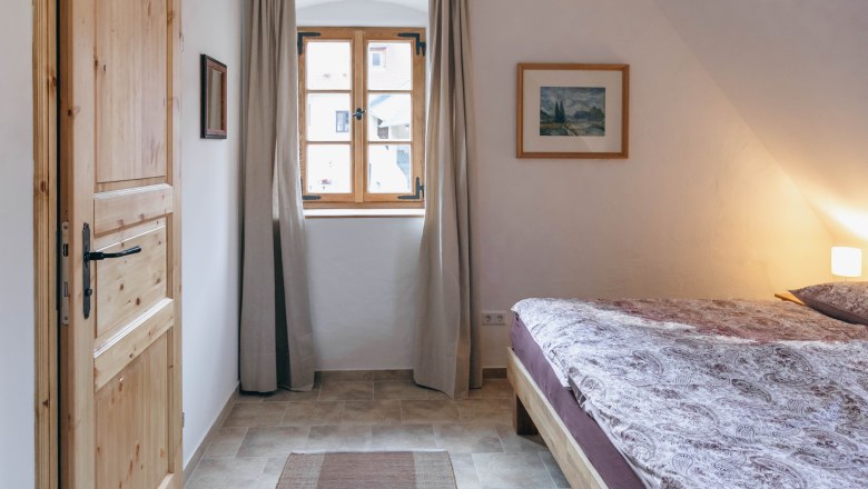 Ein gem&uuml;tliches Schlafzimmer mit Holzt&uuml;r, Fenster mit Vorh&auml;ngen, Doppelbett und Wandbild.