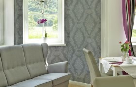 Innenraum eines stilvollen Wohnzimmers mit grauem Sofa, dekorativer Tapete, Kronleuchter und Esstisch mit Blumen.