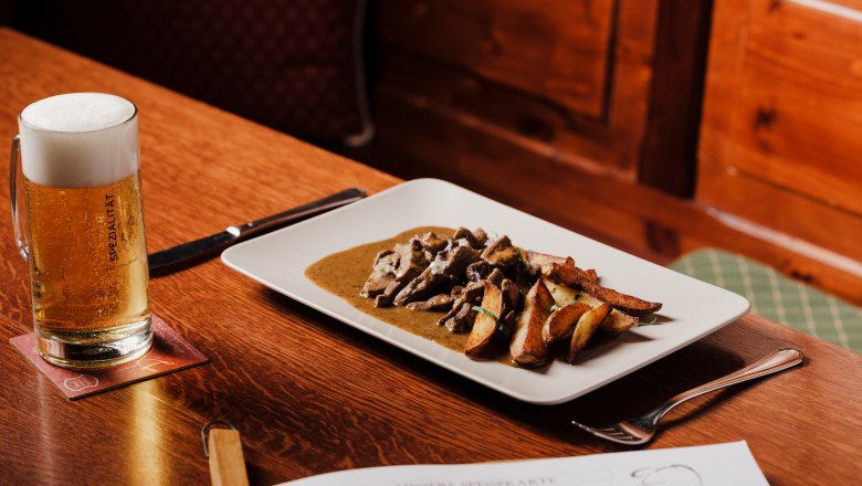 Tisch mit Bier und Teller mit Fleischgericht in rustikalem Ambiente.